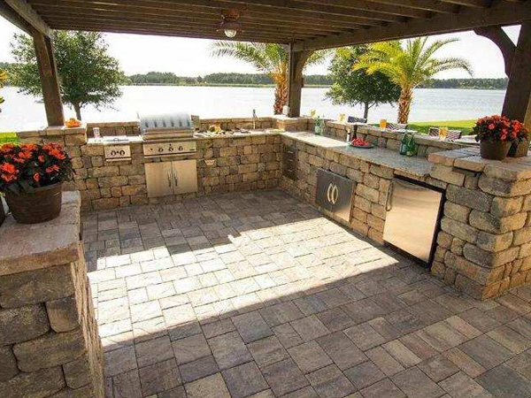 Outdoor kitchen with stainless steel appliances facing a body of water, with trees in the background.