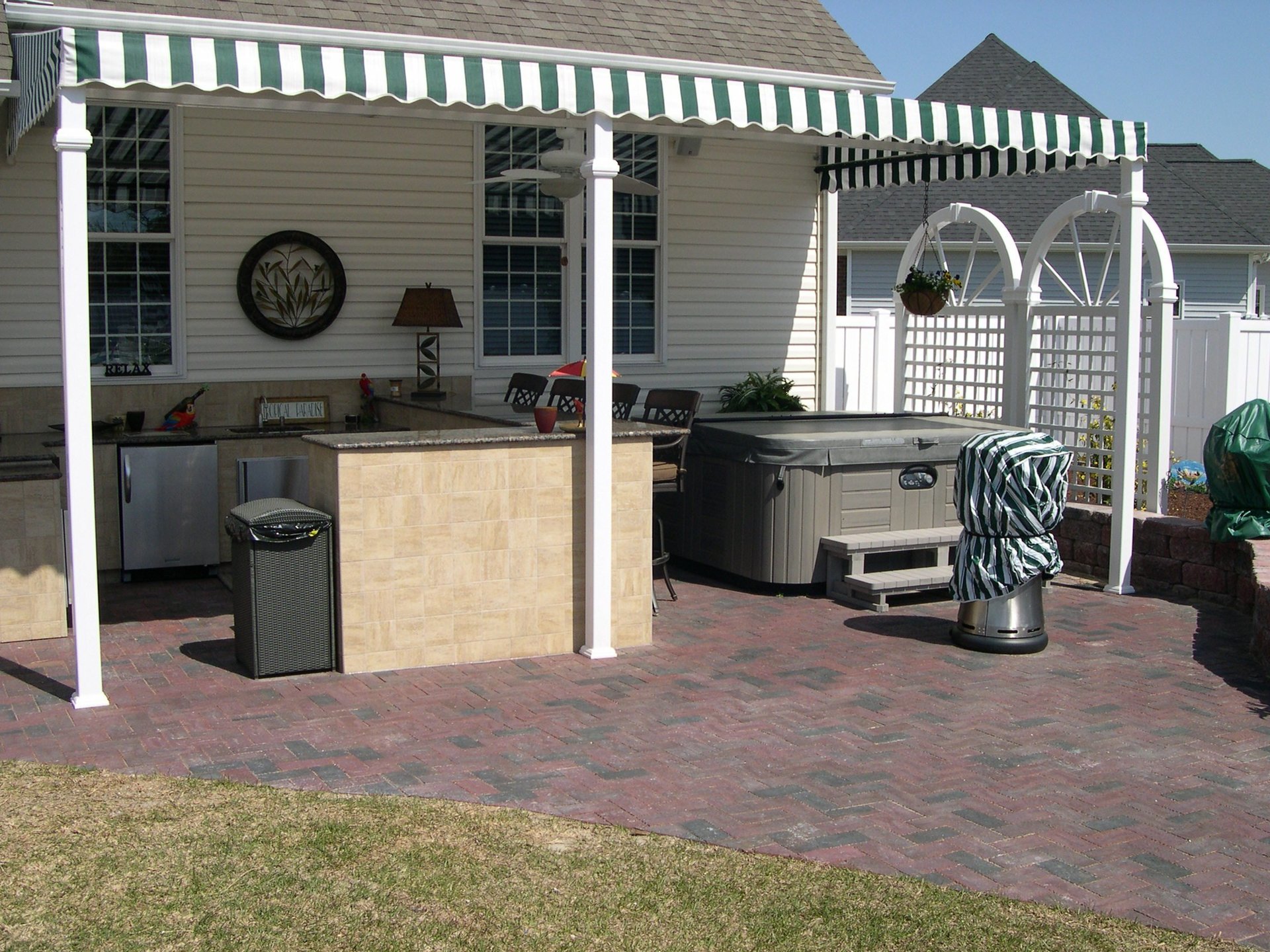 Outdoor kitchen covered by an awning featuring sink, fridge, and storage space. Adjacent to a hot-tub.